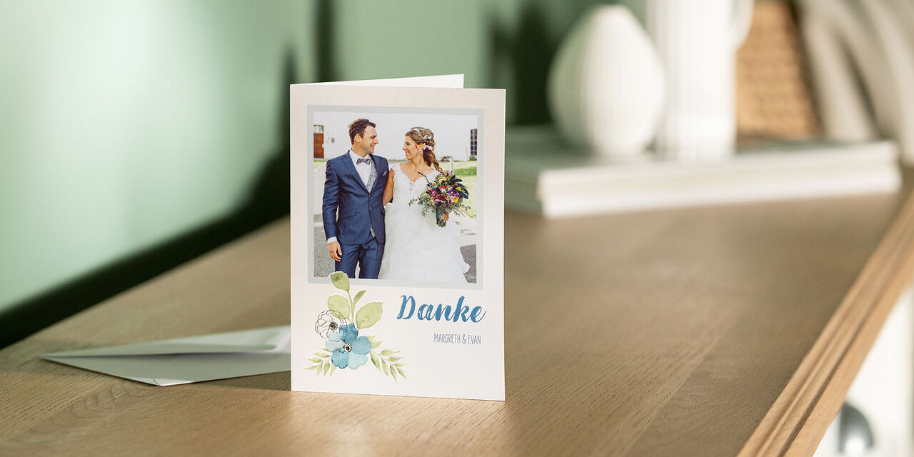 Eine Dankeskarte steht aufgeklappt auf einer Kommode. Auf der Vorderseite ist das Bild eines Brautpaares zu sehen. Darunter befindet sich die Aufschrift „Danke“ und ein florales Design. Neben der Karte liegt ein Umschlag.