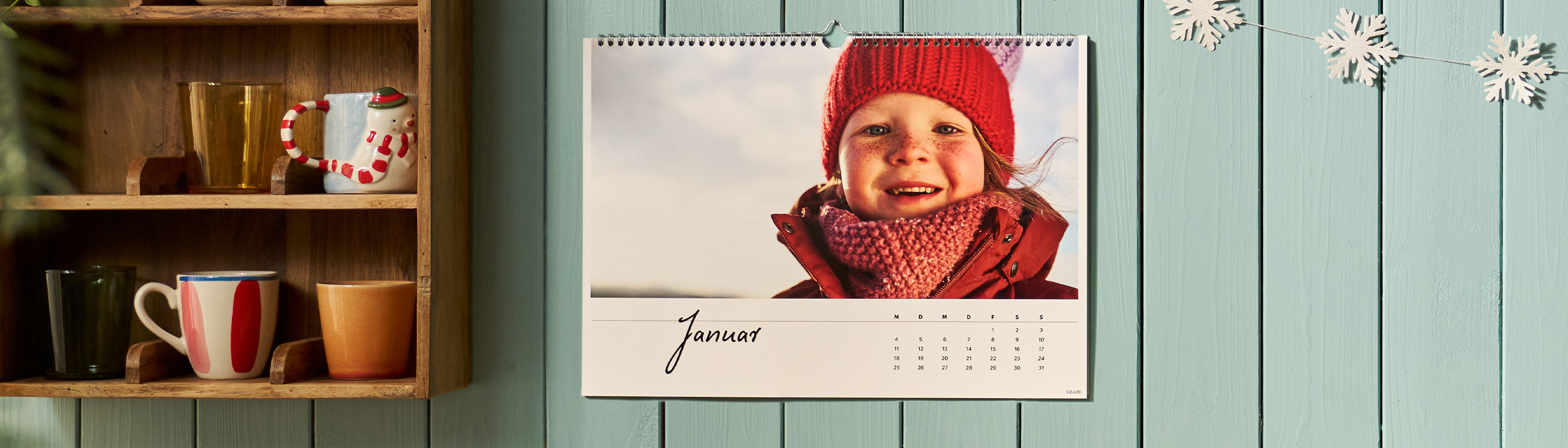 CEWE Fotokalender an türkiser Holzwand mit einem Kinderfoto und dem Monatsnamen „Januar“ neben einem Tassenregal und winterlicher Dekoration
