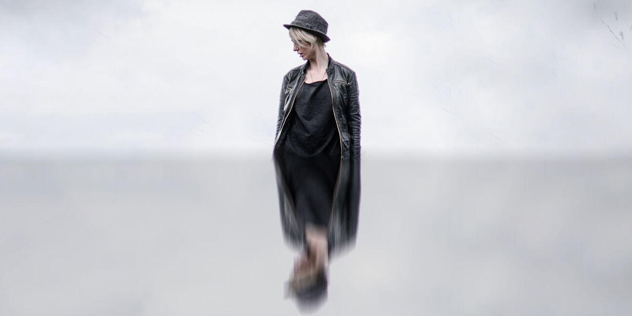 Eine Frau mit schwarzer Kleidung und Hut schaut nach unten links. Um sie herum sind nur Wolken zu sehen. Die untere Bildhälfte ist verschwommen gespiegelt.