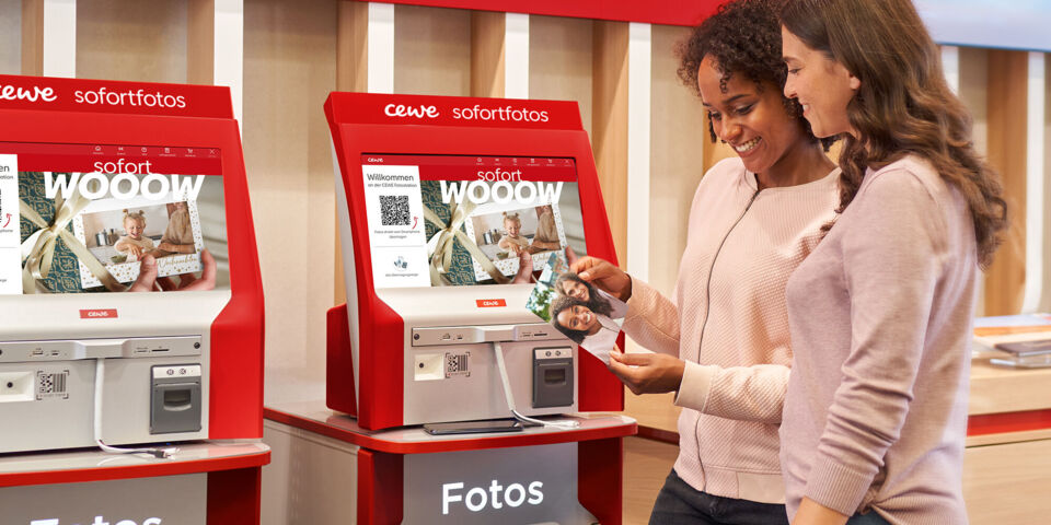 Zwei Personen nutzen rote CEWE Sofortfoto-Automaten in einer Handelspartner-Filiale.