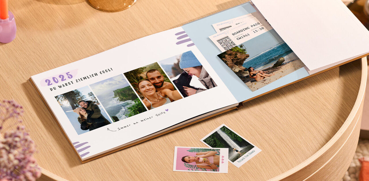 Ein offenes CEWE FOTOBUCH mit Urlaubserinnerungen und Fotoabzügen auf einem Tisch.