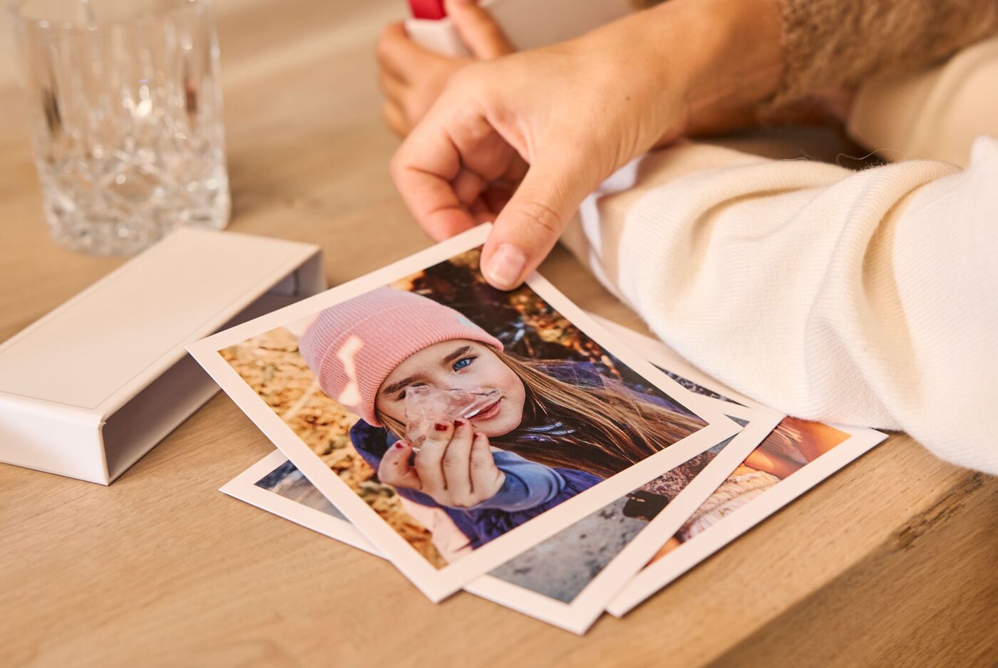 Eine Hand hält mehrere quadratische Fotoabzüge mit weißem Rahmen, die auf einem Tisch ausgebreitet liegen