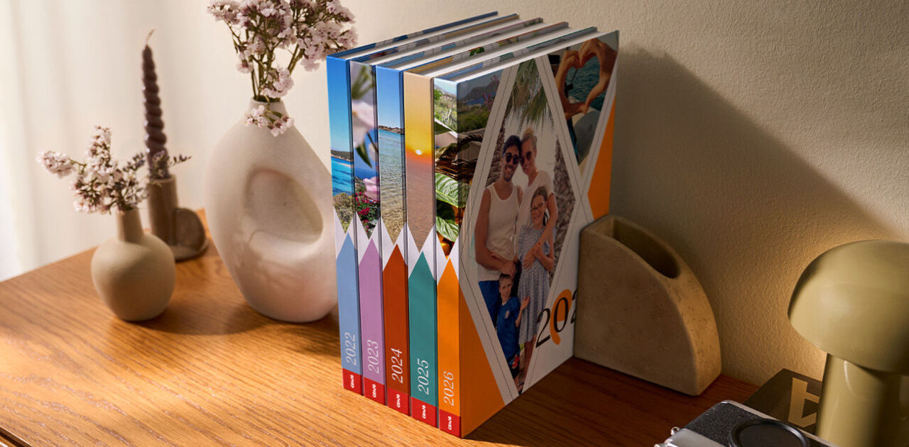 Fünf CEWE Fotobücher stehen auf einem Holztisch, umgeben von hübschen Vasen und Blumen.