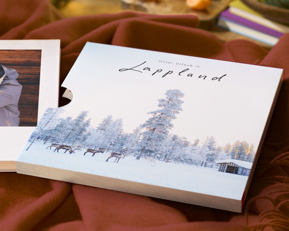 Ein CEWE FOTOBUCH mit Schuber und einer Winterlandschaft aus Lappland mit einem lächelnden Paar.