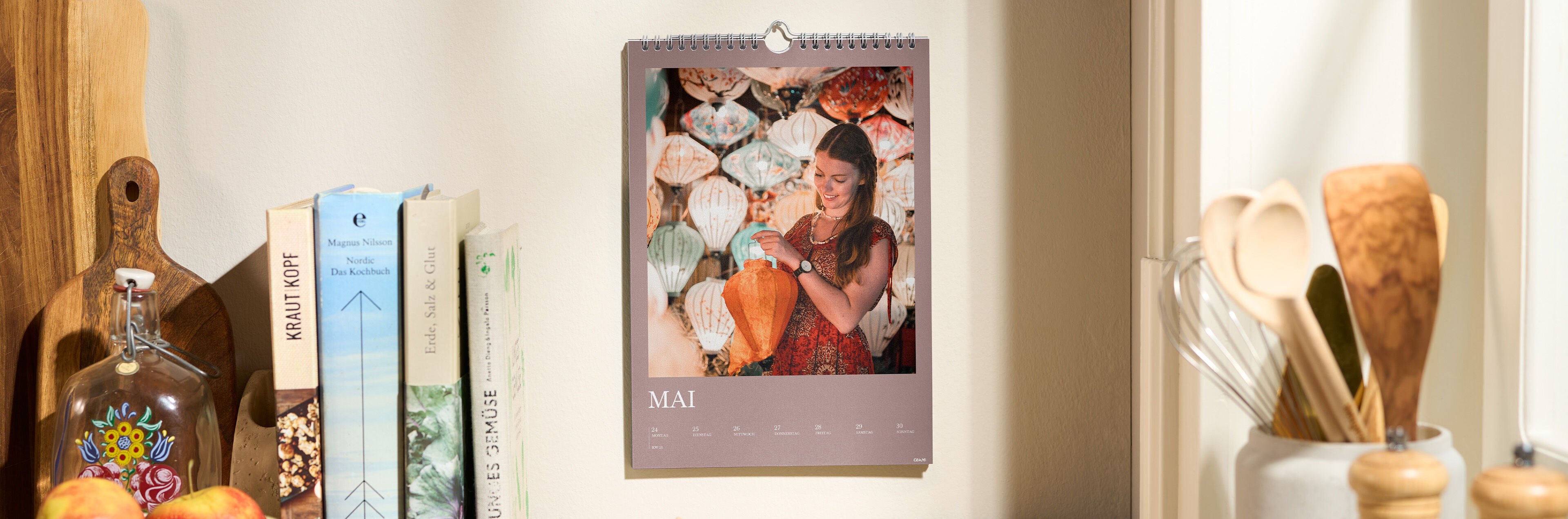 Ein CEWE Fotokalender hängt in einer gemütlichen Küche an der Wand, während eine Person lächelt.