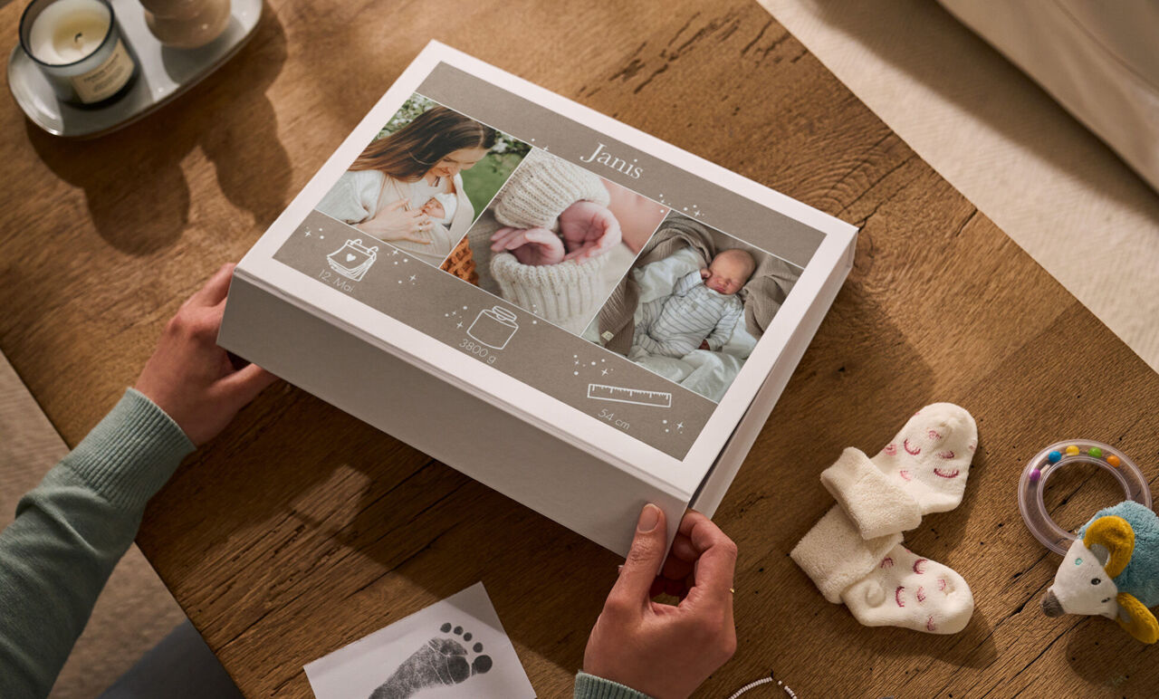 Person öffnet eine weiße Erinnerungsbox mit drei Babyfotos, dem Namen „Janis“ und kleinen Erinnerungen auf einem Holztisch.