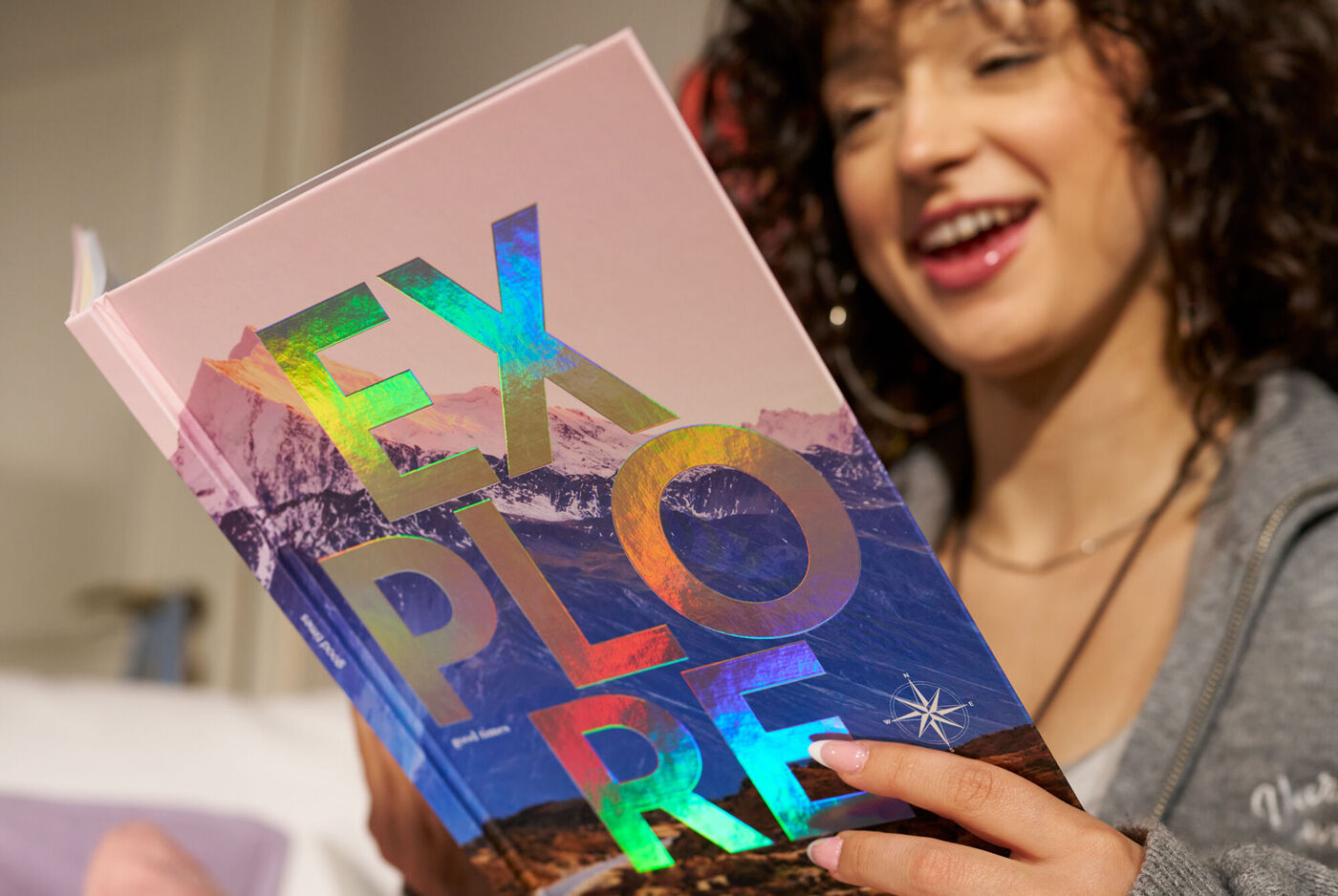 Lachende Person hält ein CEWE FOTOBUCH mit Bergmotiv und dem Schriftzug „Explore“ mit Veredelung mit holografischem Effekt