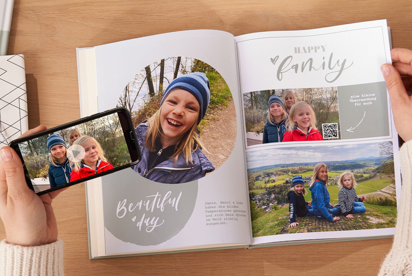 Person hält CEWE FOTOBUCH mit Familienfotos und Smartphone mit abspielbarem Video von drei Kindern.