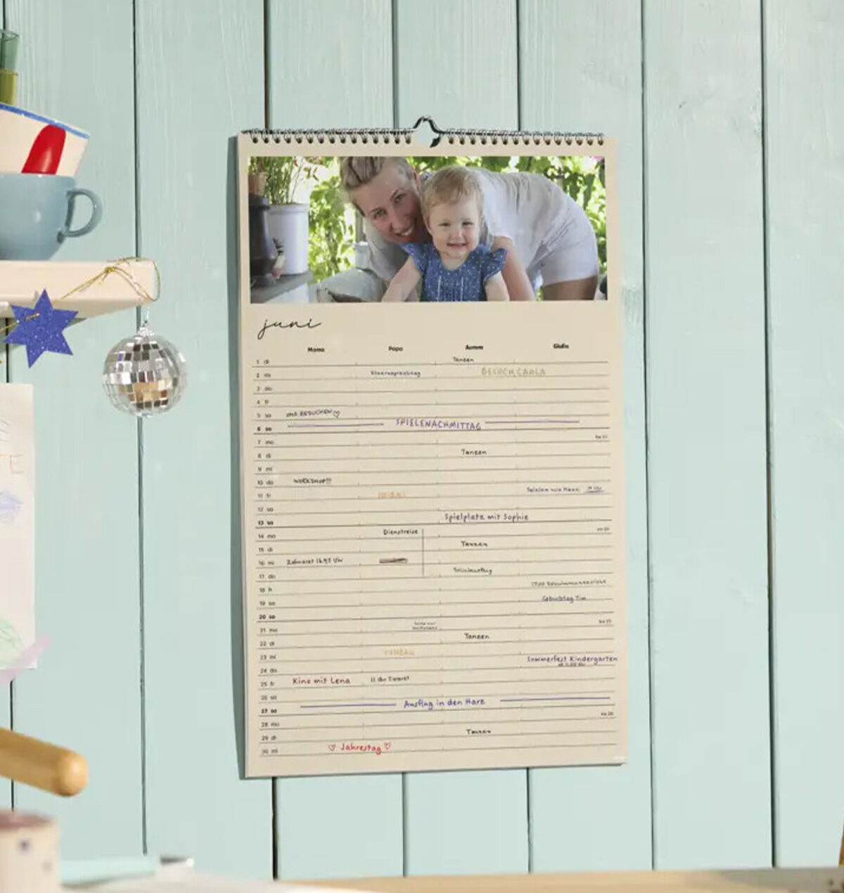 Ein CEWE Fotokalender mit einem Foto von einer lachenden Person und einem Kind hängt an einer holzvertäfelten Wand.