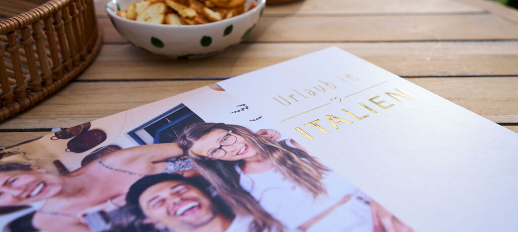 CEWE FOTOBUCH mit Schriftzug „Urlaub in ITALIEN“ mit Goldveredelung und Fotos fröhlicher Menschen auf einem Gartentisch