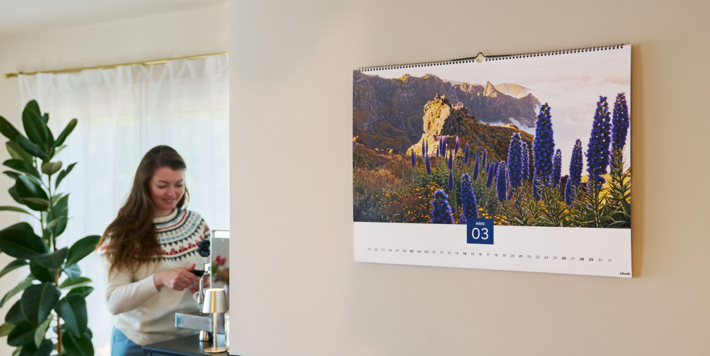In einem Wohnzimmer hängt ein großer Wandkalender, im Hintergrund steht eine Frau an einer Kaffeemaschine und bereitet etwas zu.
