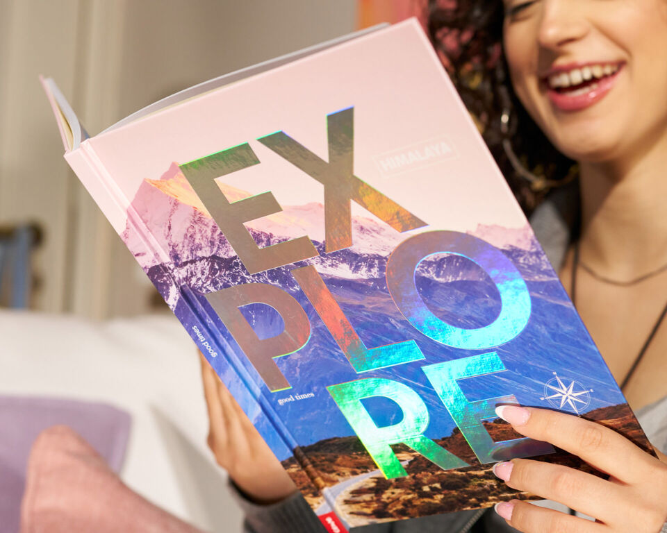 Eine Person lächelt und hält ein Buch mit dem Titel „EXPLORE“ und einer schimmernden Veredelung.vvvvvvvvvvvvvvvv