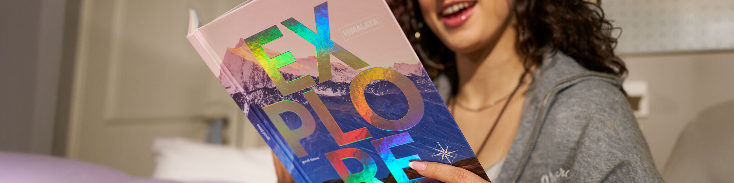 Lachende Person hält ein CEWE FOTOBUCH mit Bergmotiv und dem Schriftzug „Explore“ mit Veredelung mit holografischem Effekt
