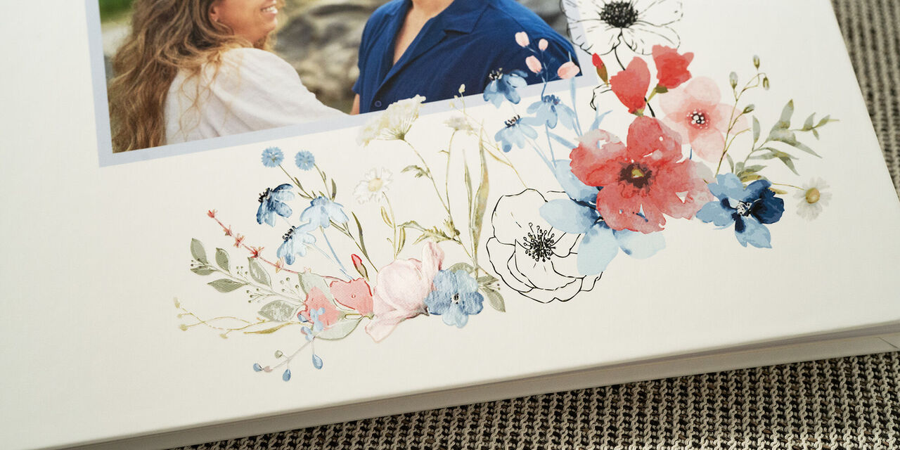 Zu sehen ist eine Nahaufnahme des Gästebuch-Covers. Es zeigt ein Bild des Hochzeitspaares. Darunter befindet sich ein florales Hochzeitsdesign mit Effektlack-Veredelung auf weißem Hintergrund.