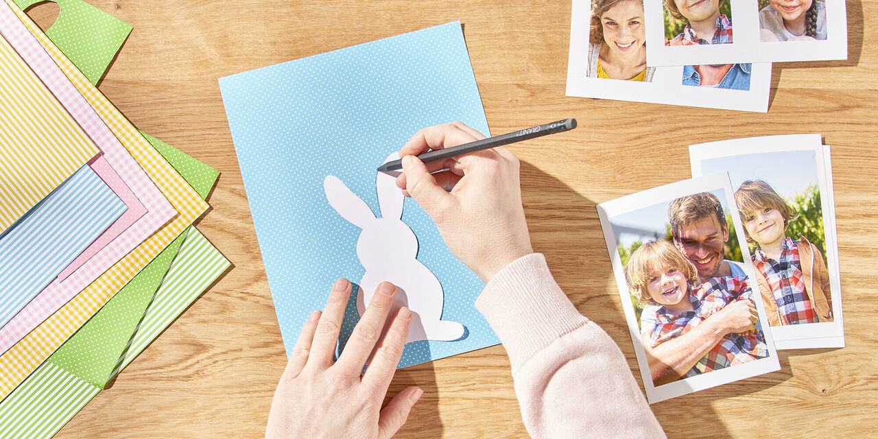 Zwei Hände zeichnen mit Hilfe der Vorlage einen Osterhasen auf das Tonpapier. Daneben liegen die ausgedruckten Sofortfotos der Familie.