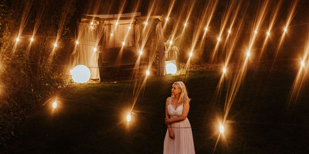 Eine Frau in einem weißen langen Kleid steht draußen in der Nacht. Um sie herum sind viele Lichtstreifen sowie im Hintergrund ein beleuchteter Pavillon.