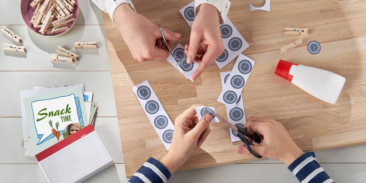 Vier Kinderhände schneiden über einer Holzplatte kleine Zifferblätter mit unterschiedlichen Uhrzeiten aus. Daneben liegen Square Prints, Klebstoff und Wäscheklammern aus Holz.