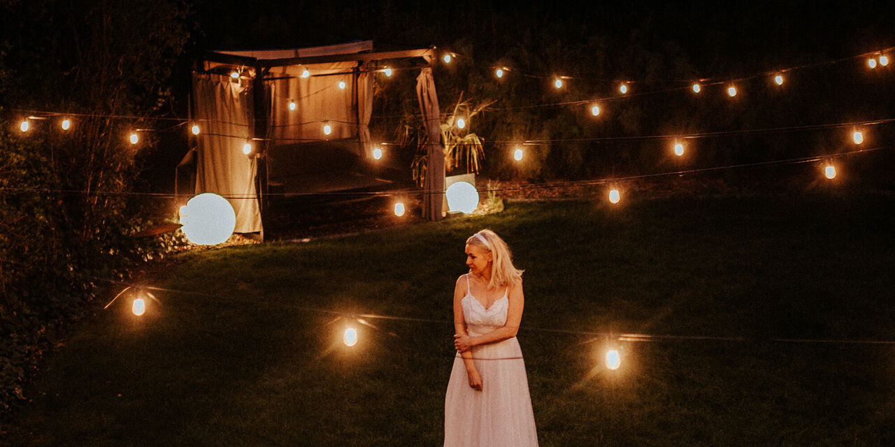 Eine Frau in einem weißen langen Kleid steht draußen in der Nacht. Um sie herum sind viele Lichter sowie im Hintergrund ein beleuchteter Pavillon.