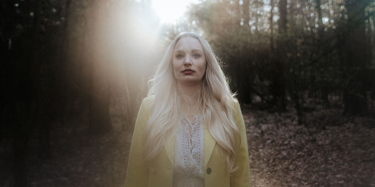 Eine blonde Frau mit einem gelben Mantel steht im Wald. Von hinten scheinen Sonnenstrahlen durch die Bäume auf sie. Insgesamt ist die Umgebung und das Licht verschwommen.
