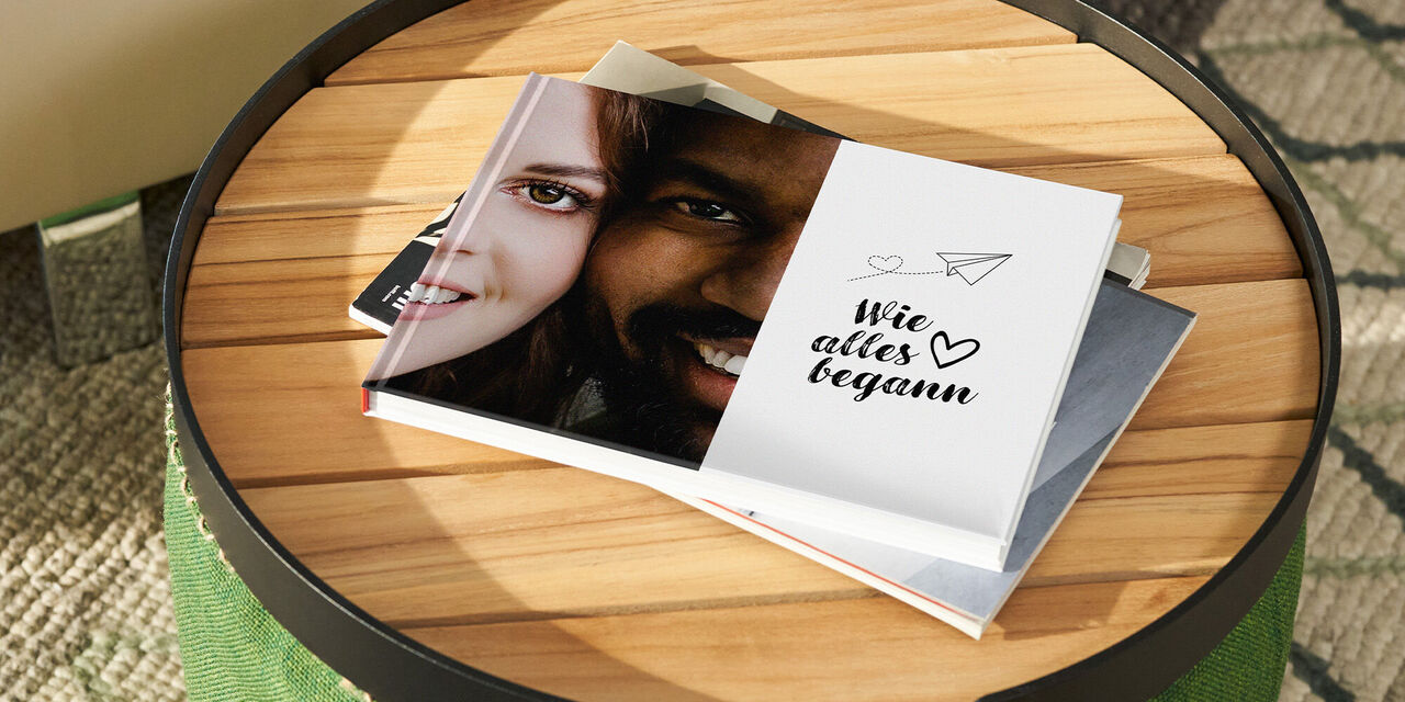 Ein CEWE FOTOBUCH liegt zugeklappt auf einem Holztisch. Auf dem Cover ist eine Nahaufnahme eines Paares zu sehen und der Text "Wie alles begann".