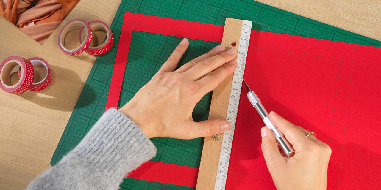 Zwei Hände halten ein Lineal und einen Stift. Eines der so abzumessenden vier Fenster wurde bereits in den roten Pappkarton geschnitten.