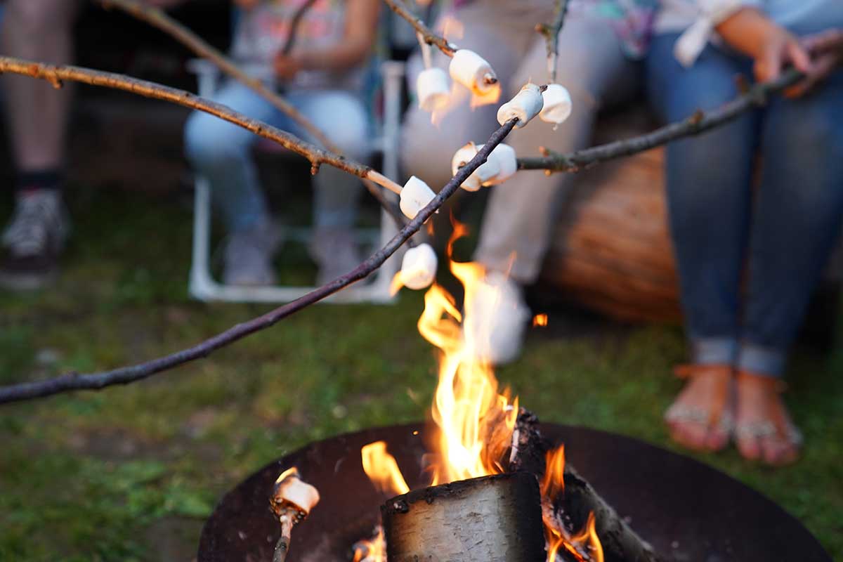 Familie Fischer kreuzt gemeinsam Stöcker über dem Lagerfeuer. Sie bereiten Marshmellows zu.