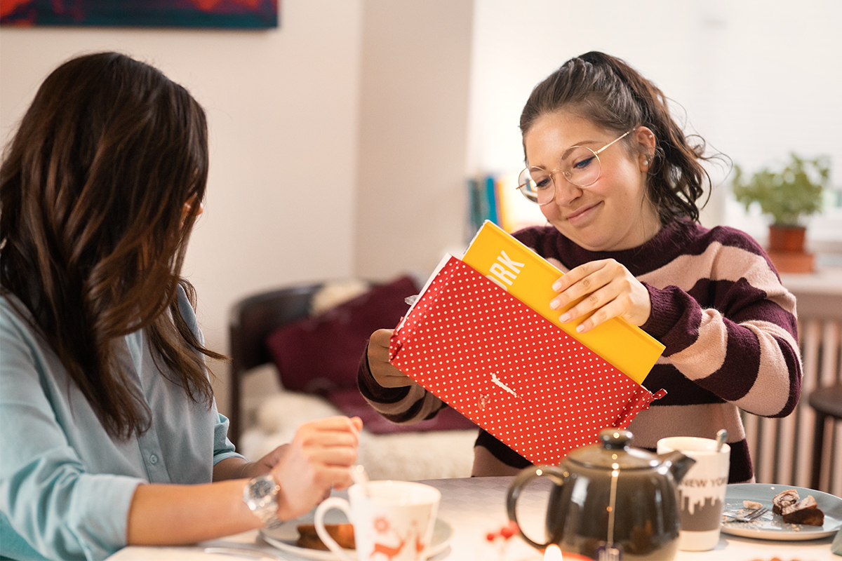 Lilli packt am Tisch das CEWE FOTOBUCH aus einer Geschenkverpackung aus.