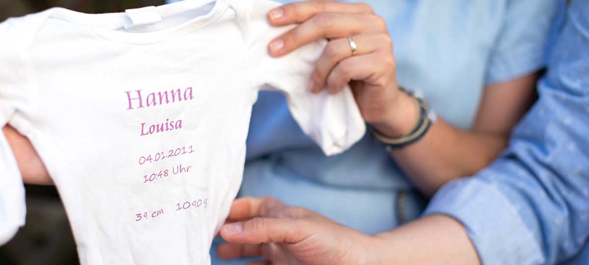 Tina Kneitz und Melanie Kofler halten gemeinsam einen Babystrampler in den Händen.